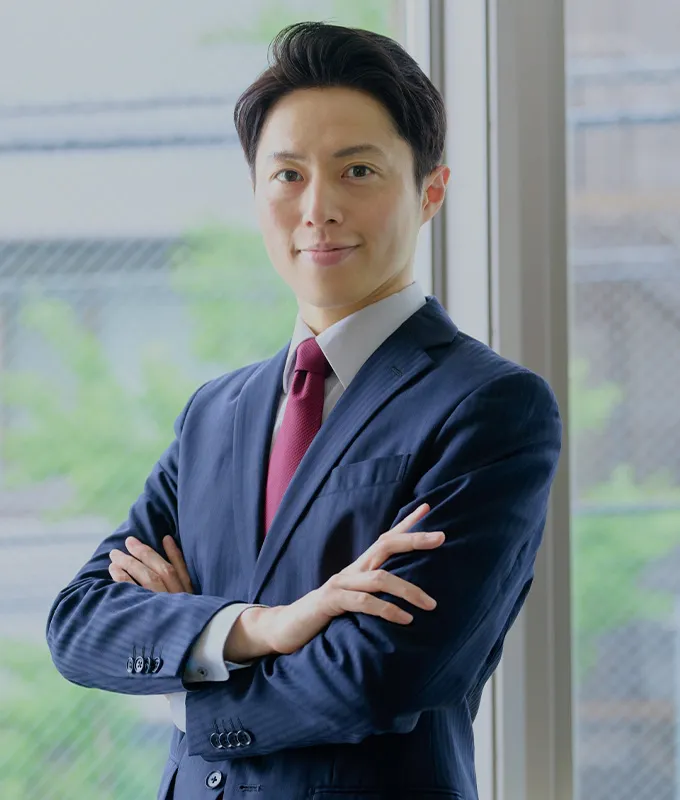 オフィスの窓際で腕を組んでこちらを見るスーツ姿の男性